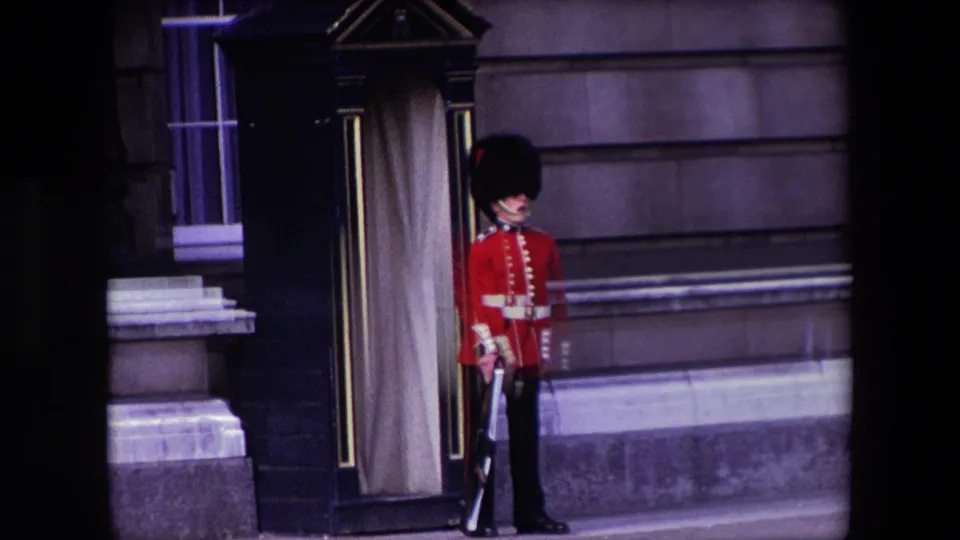 A Queen's Guard stands in red ceremonial dress with a rifle outside a stone London building, framed by the dark edges of the 8mm film scan.