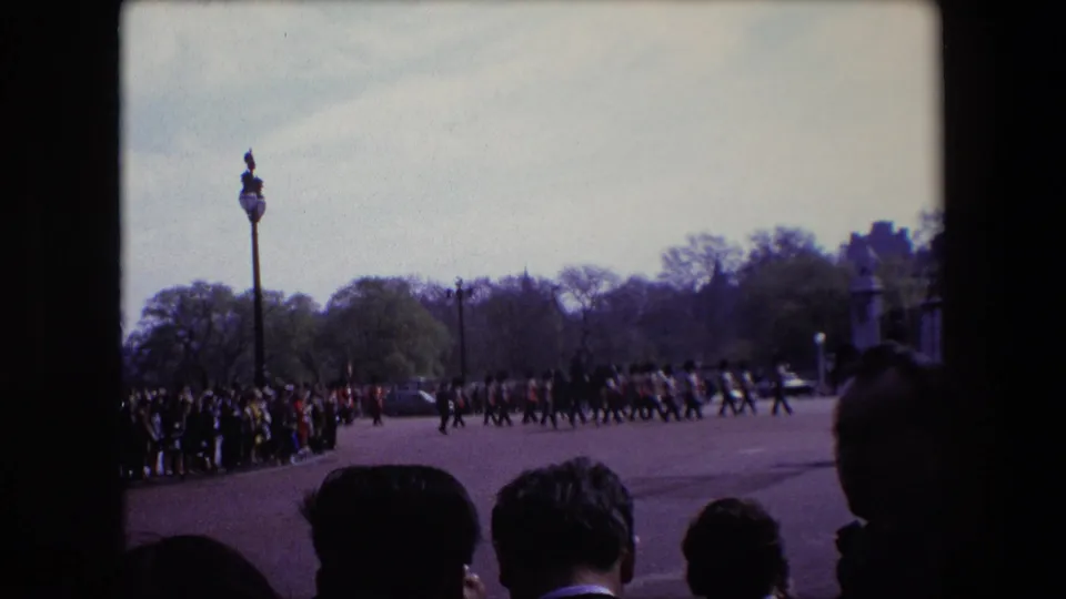 In London, England in 1967, spectators line the edge of a broad paved route while uniformed soldiers march in formation past trees and lampposts.