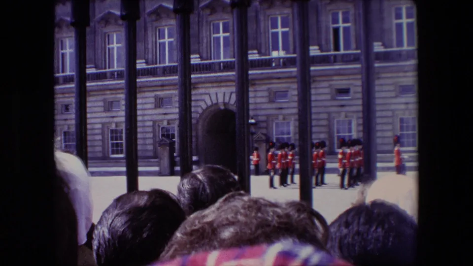 1967 London home-movie frame looking through iron railings toward red-uniformed guards near a large formal building, with crowd heads at the bottom of frame.