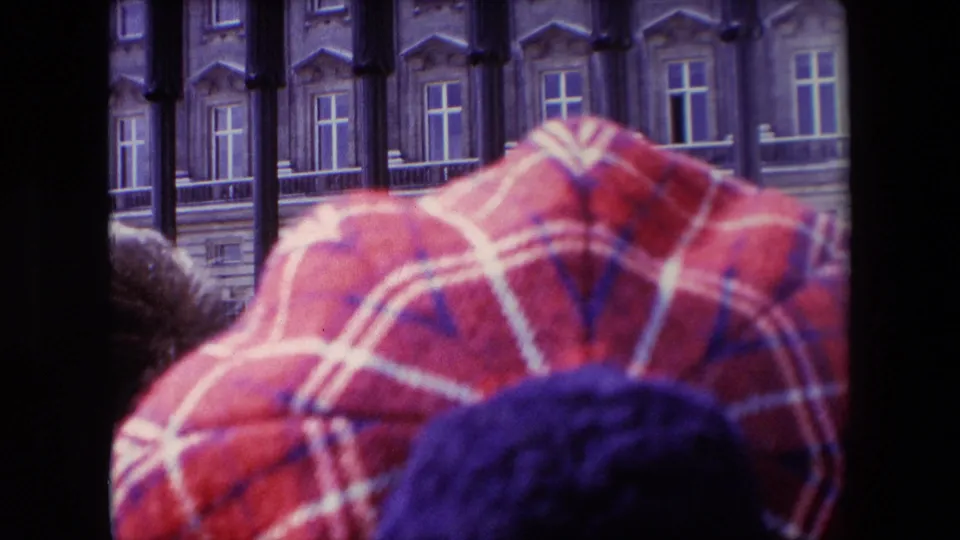 1967 London home-movie frame from within a spectator crowd, with a red patterned cap in the foreground and guards visible beyond iron railings near a formal building.