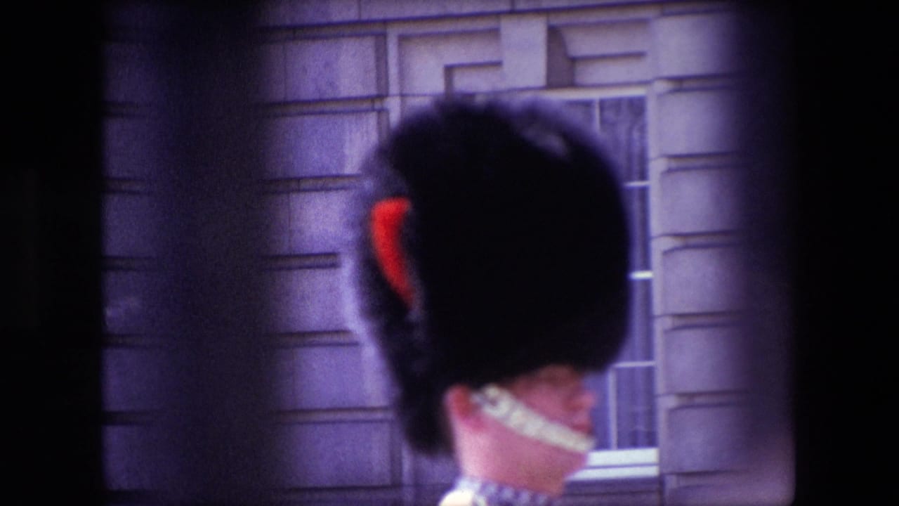 1967 London home-movie frame showing the back of a Royal Guard's tall black hat with red plume and white chin strap against a stone building facade.