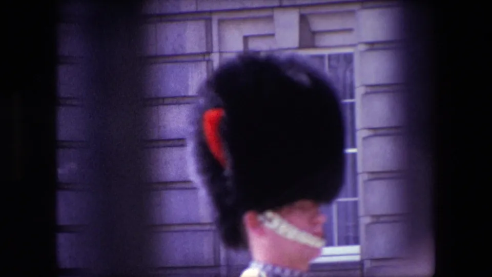 1967 London home-movie frame showing the back of a Royal Guard's tall black hat with red plume and white chin strap against a stone building facade.