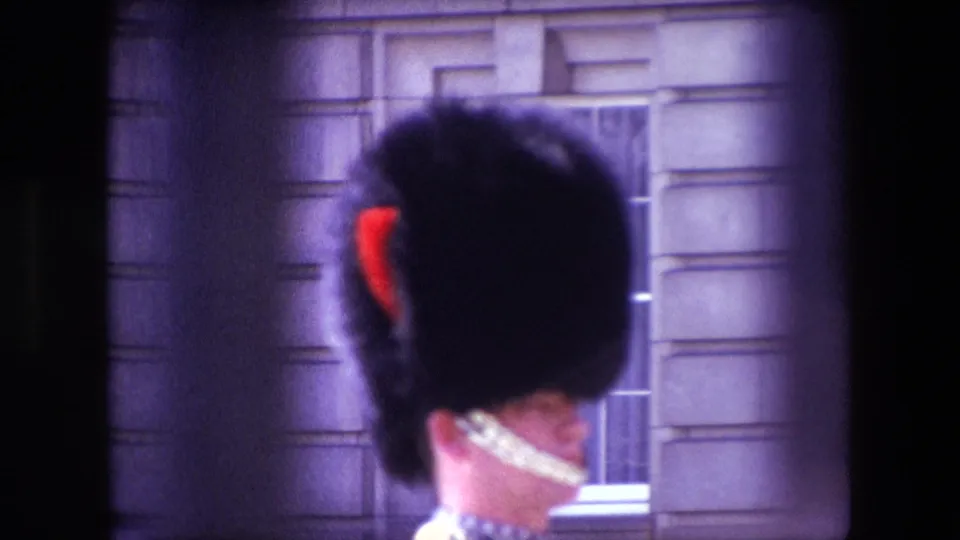 1967 London frame showing a Royal Guard from behind, with a red plume and white chin strap visible against a formal stone facade and window.