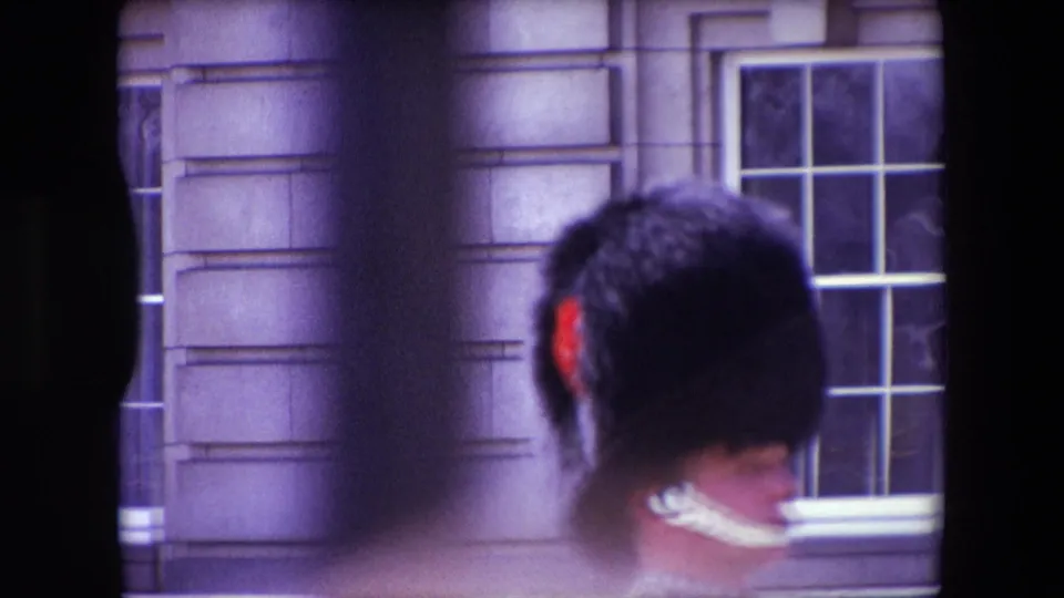 Soft 8mm frame from London in 1967 showing the rear of a Royal Guard with a bearskin-style hat and white chin strap near a stone building window.