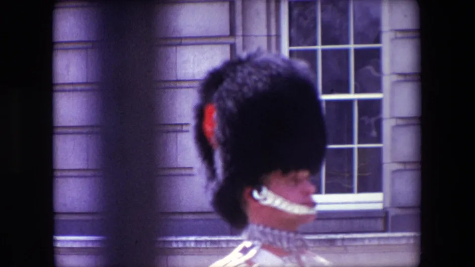 1967 London home-movie still showing a ceremonial Royal Guard from behind, with red plume, white chin strap, and stone-window background visible through soft color-shifted film.