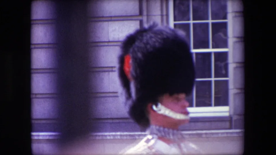 1967 London home-movie frame showing a Queen's Guard in profile near a stone facade and multi-pane window, with soft focus and visible film borders.