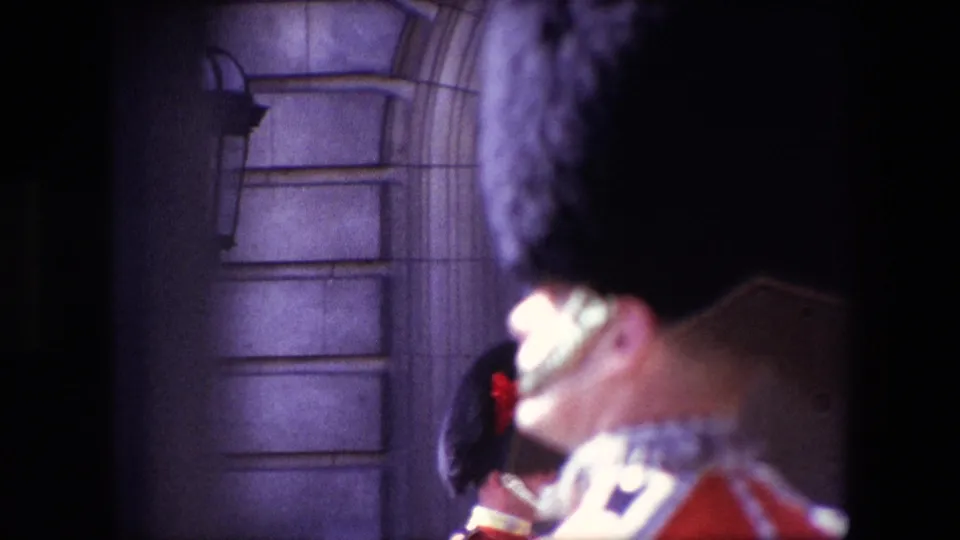 1967 London home-movie frame with a close, partially cropped view of a Queen's Guard in ceremonial uniform near stone architecture.