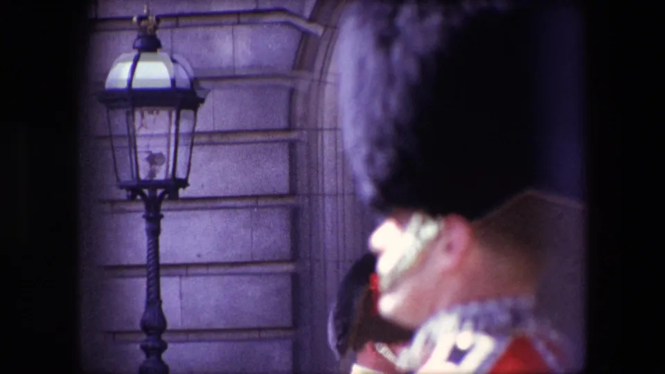 1967 London home-movie frame showing a Queen's Guard in profile near a traditional exterior lamp and stone facade during ceremonial sightseeing footage.