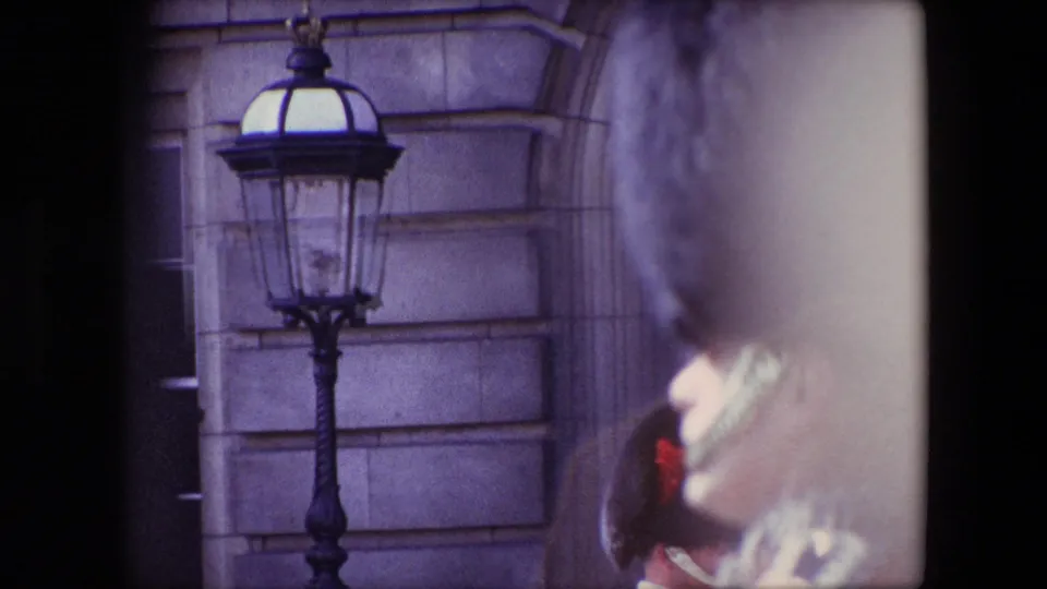 1967 London frame with a black lamp fixed before pale stone architecture while a ceremonial guard in red is partially visible and out of focus at the right.