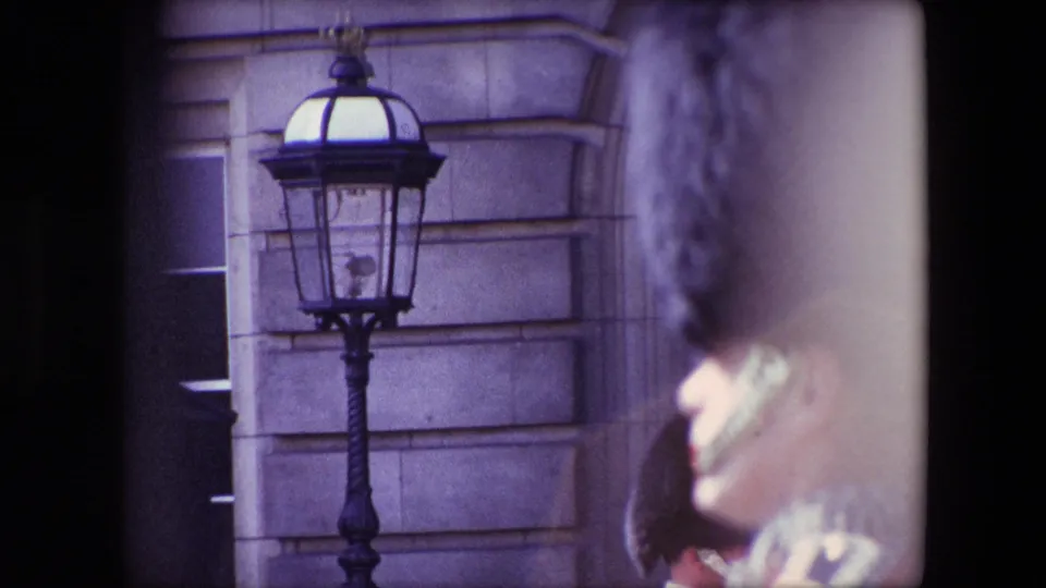 1967 London palace guard footage with a black lamp and stone facade behind a blurred red-coated guard wearing a tall black hat.