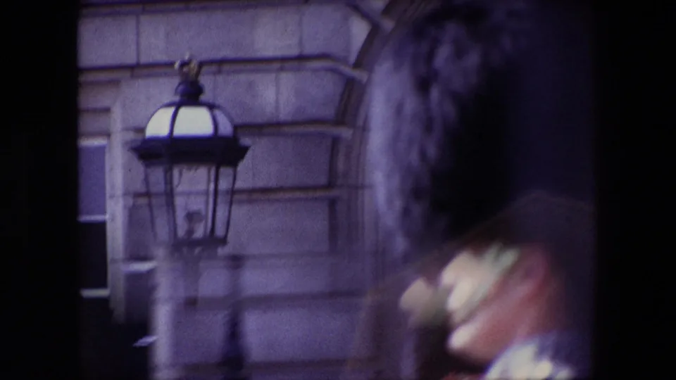 Final 1967 London frame showing a soft, handheld view of a red-uniformed palace guard moving past a black lamp and stone architectural background.