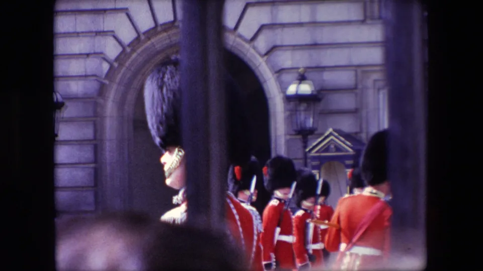 1967 Buckingham Palace home-movie frame with one guard prominent behind vertical bars and additional Queen's Guard soldiers assembled beyond him.