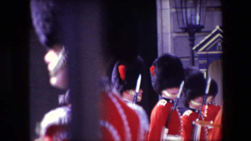 Close 8mm frame from 1967 London showing Queen's Guard soldiers in red tunics and bearskin hats near Buckingham Palace architectural details.