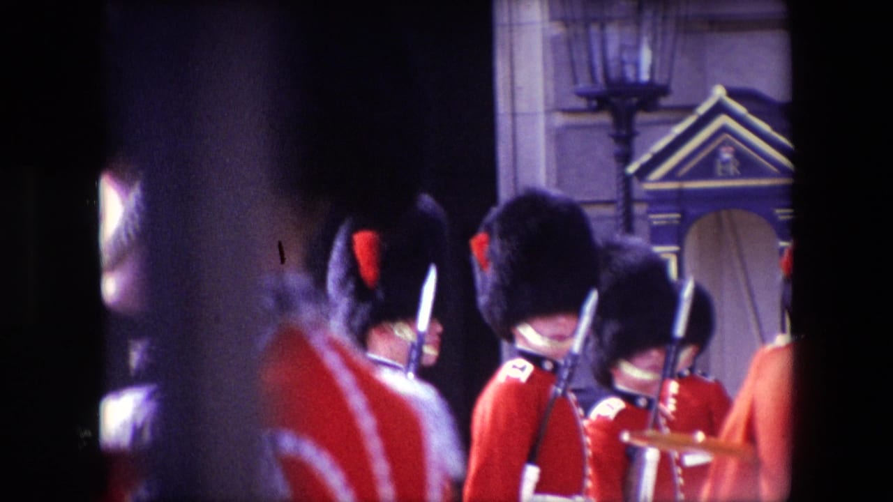 1967 London home-movie frame showing British Royal Guards in red tunics and bearskin hats, partially obscured by nearby spectators and dark foreground shapes.