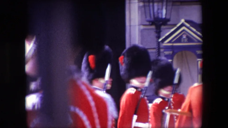 1967 London frame of Royal Guards in red coats and bearskin hats standing with rifles near a stone facade and decorative guard structure, filmed from behind spectators.