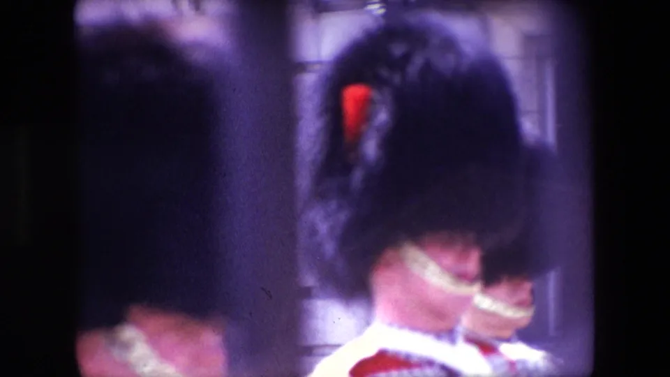 Soft 8mm frame from 1967 London showing a close obstructed view of a Royal Guard's bearskin hat and red uniform, with another ceremonial guard partially visible behind.