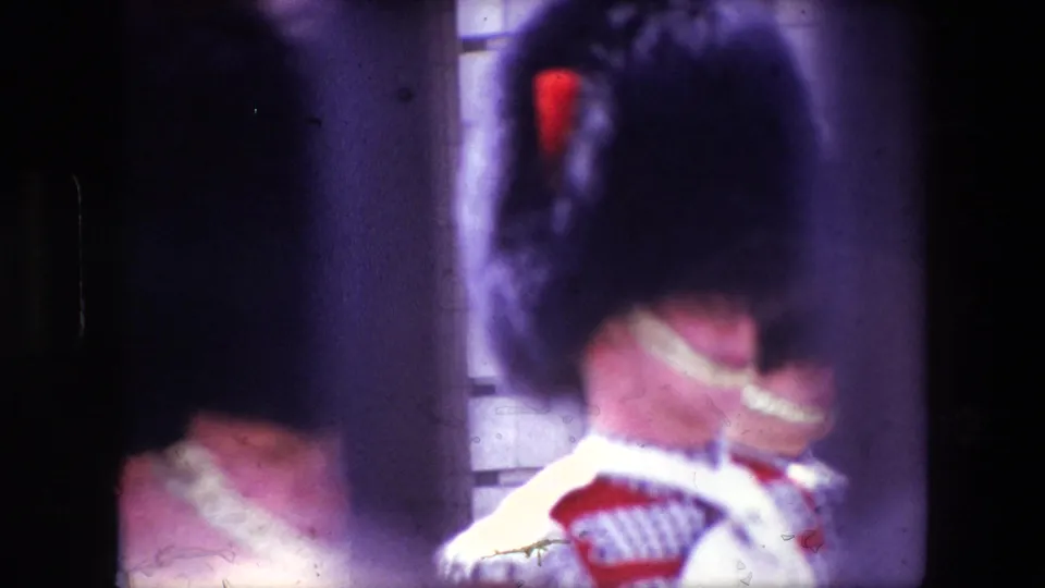 1967 London home-movie frame with a blurred close view of Royal Guards in bearskin hats and red dress uniforms, marked by purple color fading and surface scratches.