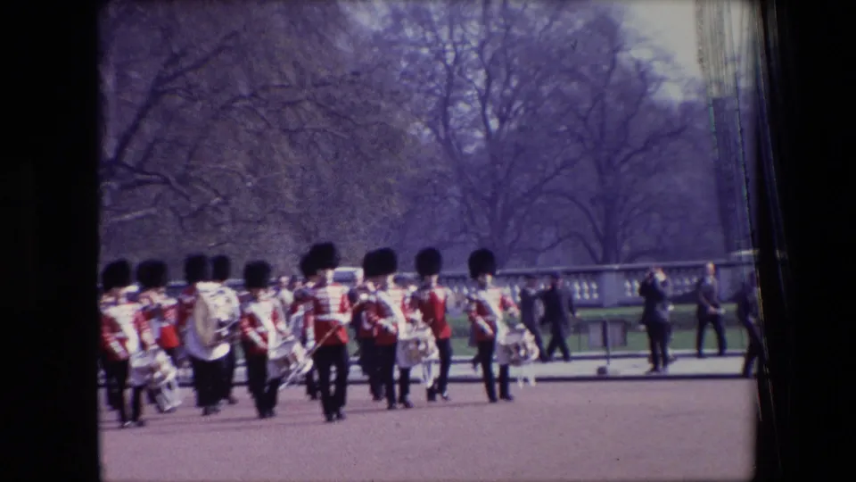 Red-coated soldiers in tall black hats march with drums and band instruments across open grounds in the supplied 1967 London ceremonial parade footage.