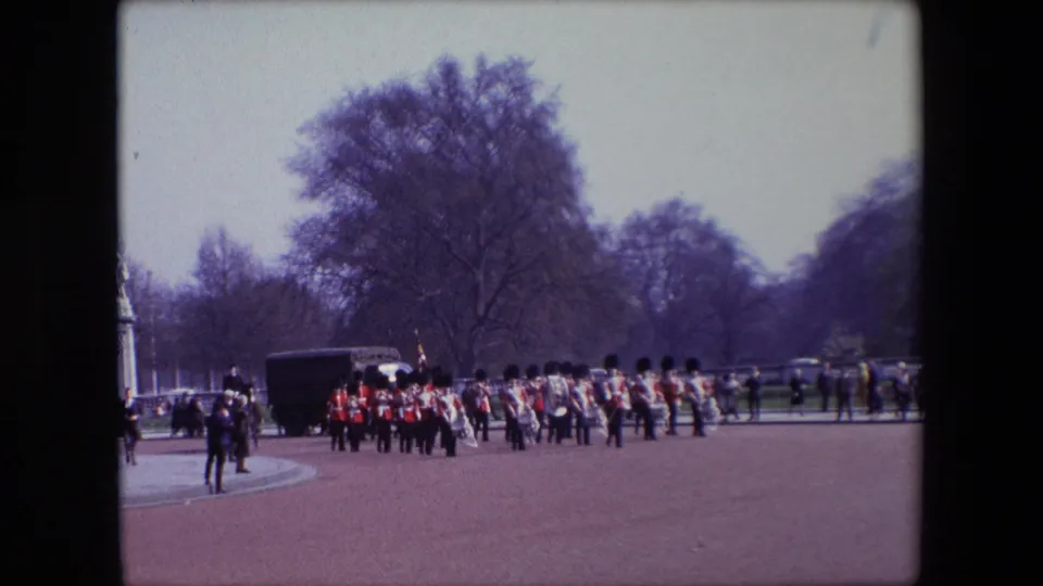 A wide London parade-ground view shows a ceremonial group of red-coated soldiers marching in formation, with trees, spectators, and a dark vehicle or structure in the background.