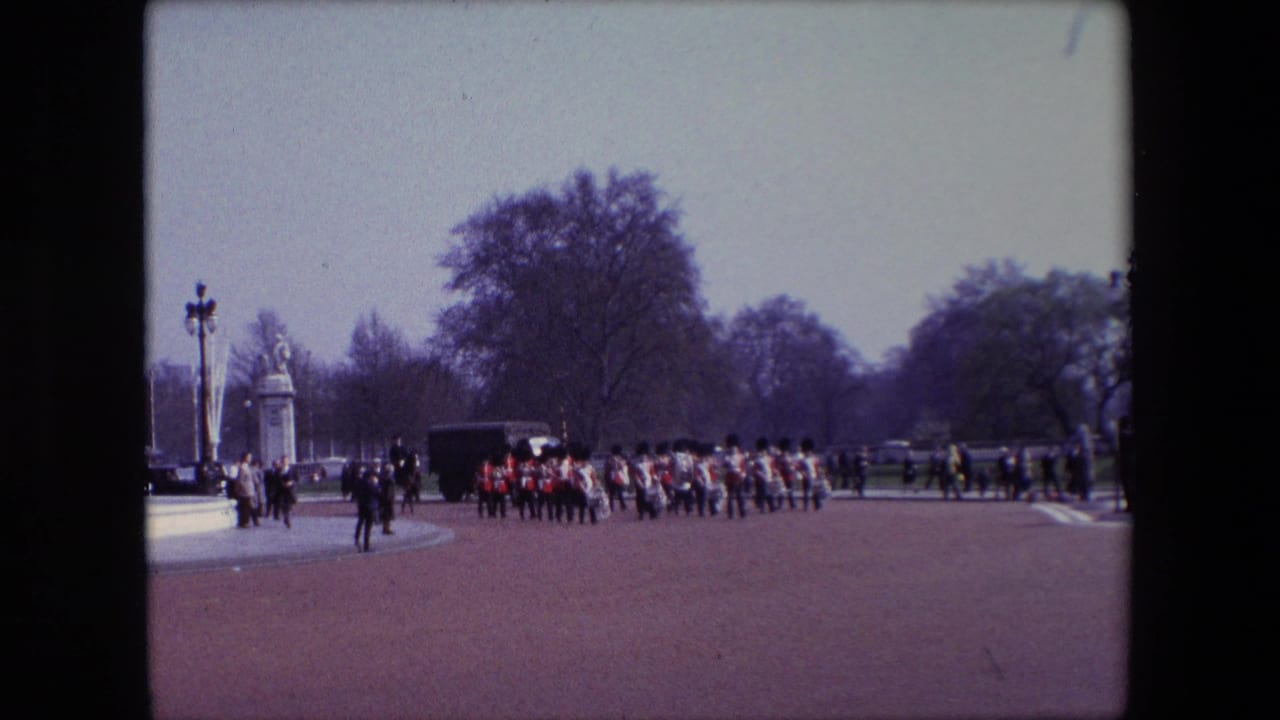 1967 London home-movie frame showing British Guards in red tunics and dark hats marching in formation across a wide parade ground, with spectators, gate structures, lampposts, and trees in the background.