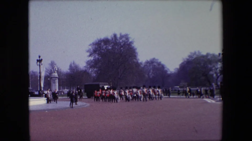 1967 London home-movie frame showing British Guards in red tunics and dark hats marching in formation across a wide parade ground, with spectators, gate structures, lampposts, and trees in the background.