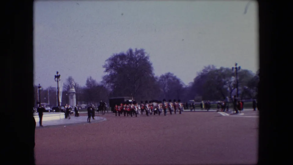 1967 London frame of British Guards moving in synchronized lines across an open ceremonial space, with distant onlookers, a dark vehicle or carriage-like form, trees, and stone gate structures visible behind them.