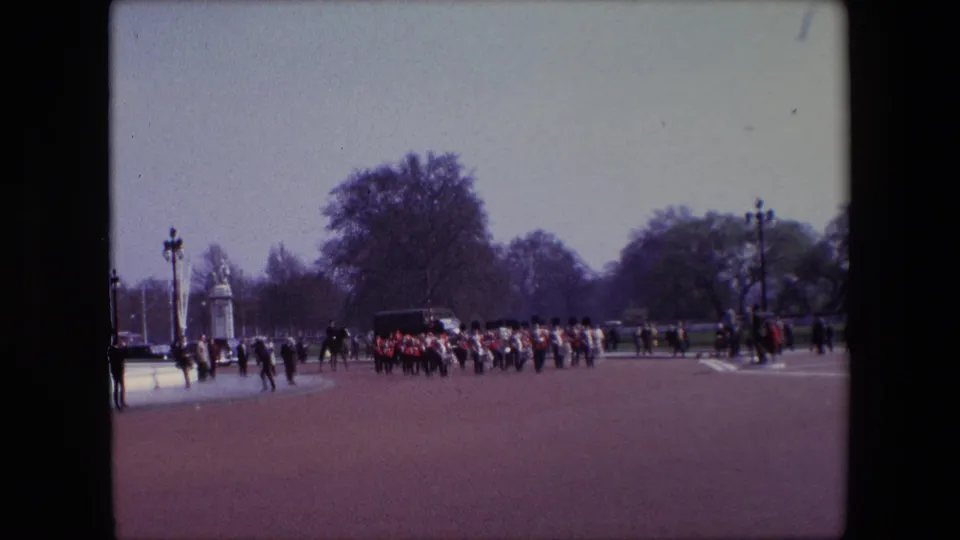 1967 London home-movie still with Guards in red uniforms marching across the paved ground, while dark-clad parade figures or mounted participants and spectators stand near the left side of the scene.