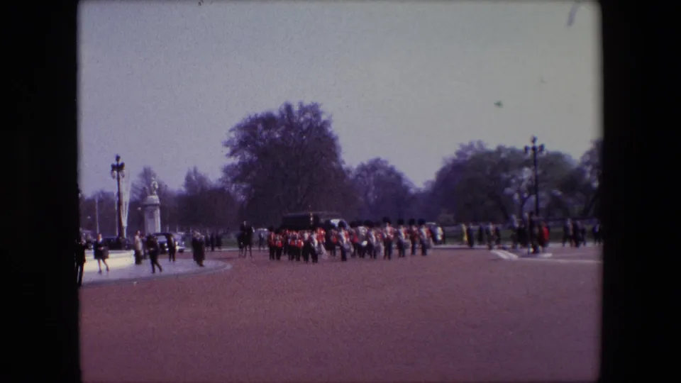Final 1967 London frame showing British Guards still moving in formation across the reddish paved forecourt, with distant spectators, lampposts, trees, and stone gate features in the background.