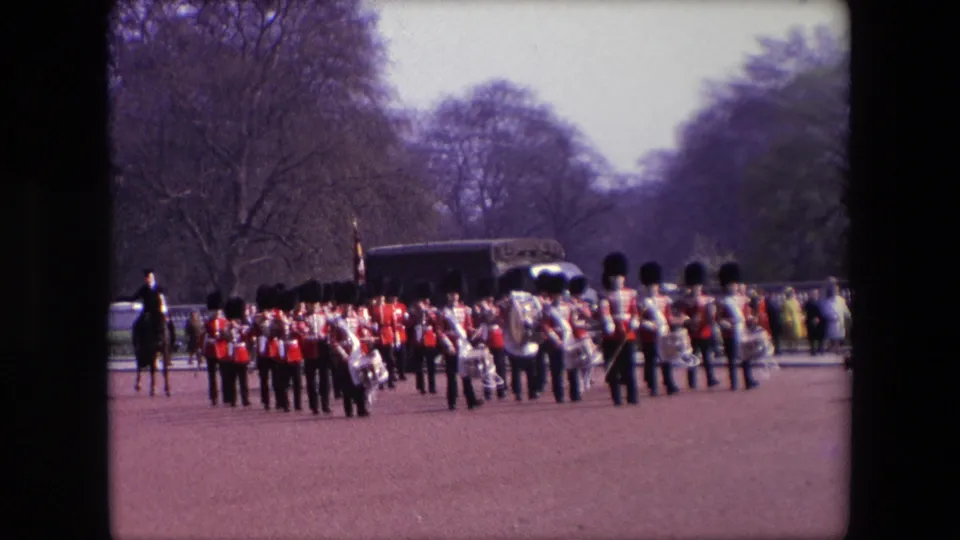The marching formation comes closer, showing guards in red tunics, band members carrying drums and instruments, a mounted officer to the left, and a dark vehicle parked behind the procession.