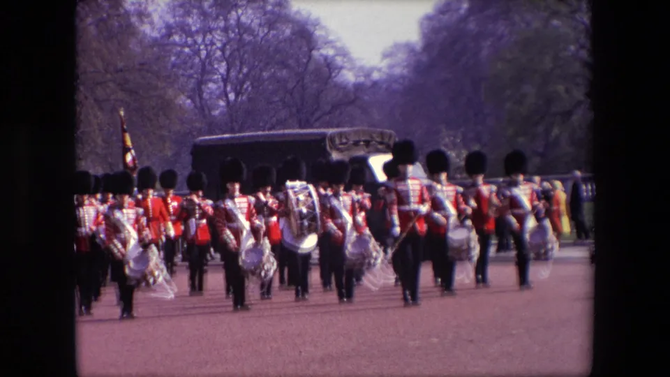 A closer 8mm frame shows British guards and band members marching toward the camera in red tunics and black bearskin hats, with drums, instruments, a dark vehicle, and spectators in the background.
