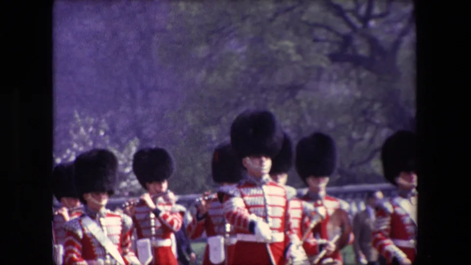 Uniformed musicians face toward the camera in a soft 8mm frame, with tall black hats, red tunics, straps, and instruments visible against trees and street background.