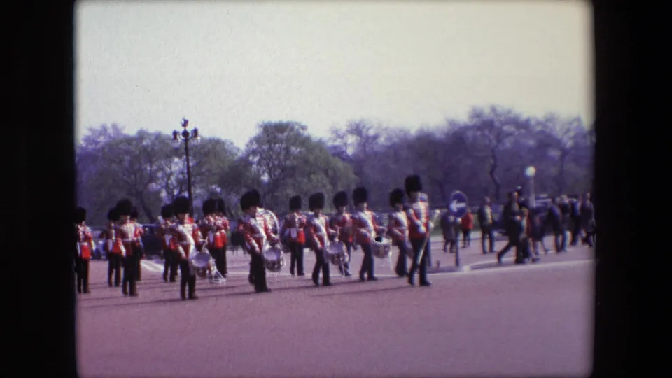 A wide 1967 London street view shows the marching group spread across the road, with uniformed musicians, pedestrians or spectators, trees, and street fixtures in the background.