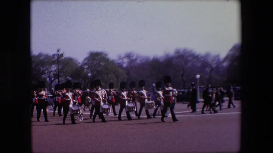 In London archival home-movie footage from the supplied 1967 metadata, uniformed band members in red jackets and tall dark hats march across a broad paved area with drums and brass instruments.