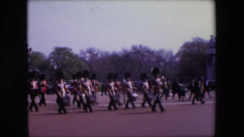 A line of uniformed musicians in red jackets and dark hats moves through the London parade scene, with drummers and brass players visible against trees and pedestrians.