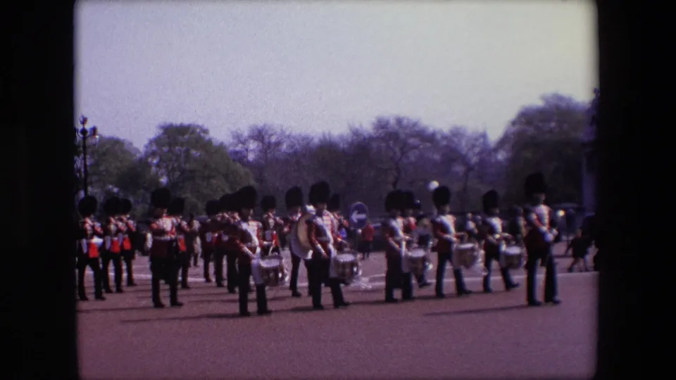 The home-movie frame shows the band partly paused or changing formation, with multiple drummers and red-uniformed musicians spread across the open London parade route.