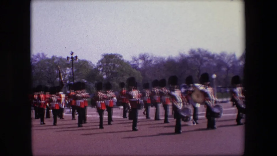Uniformed musicians in red jackets gather across the broad London parade route, with a drummer prominent near the right edge and trees and spectators in the distance.