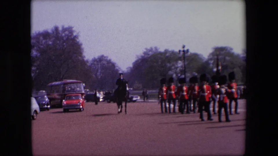 In 1967 London, guards in red uniforms stand or move in formation on a broad road with period traffic, including a red bus, visible to the side.