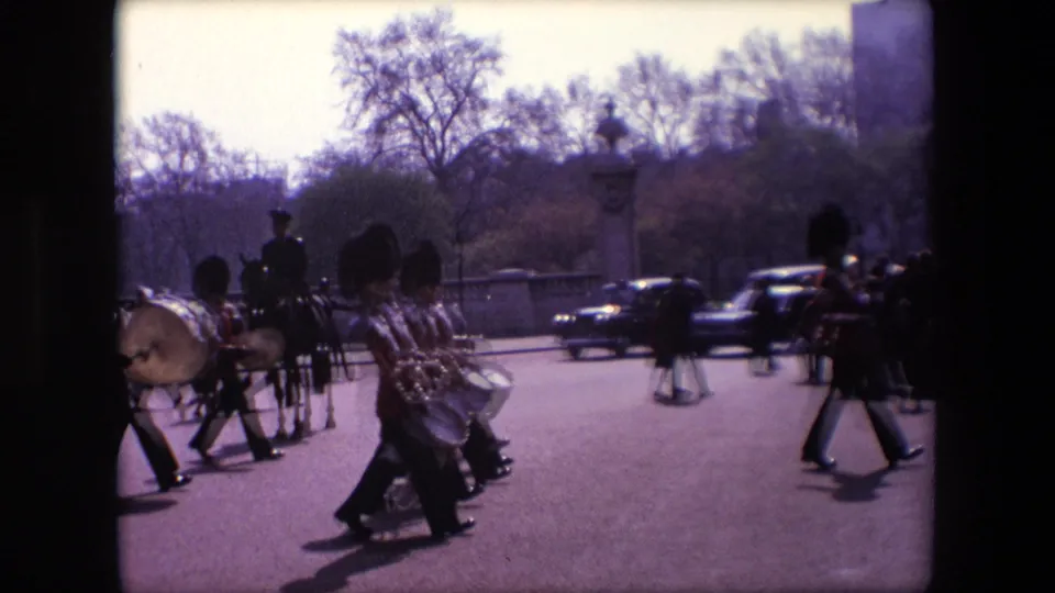 In 1967 London, ceremonial guards and band members carrying drums move along a broad tree-lined road with cars passing in the background.