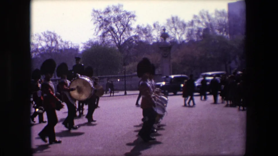 1967 London home-movie frame showing Queen's Guards in red ceremonial uniforms and tall dark hats marching with drums past spectators and cars.