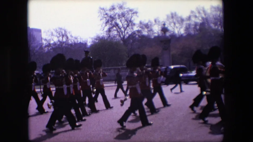 1967 London 8mm frame showing a close roadside view of Queen's Guards in red uniforms and bearskin-style hats marching with brass instruments.