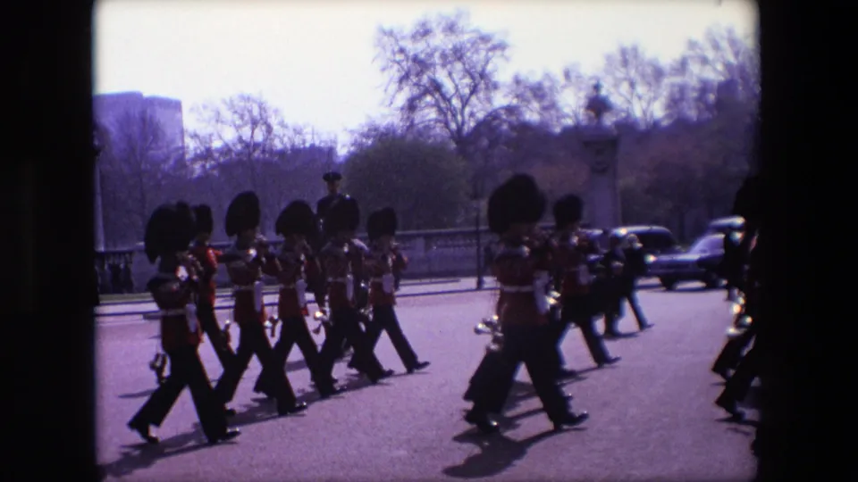 1967 London parade frame with ceremonial guards in formation, a drummer at right, brass players at center, and spectators and cars along the street.