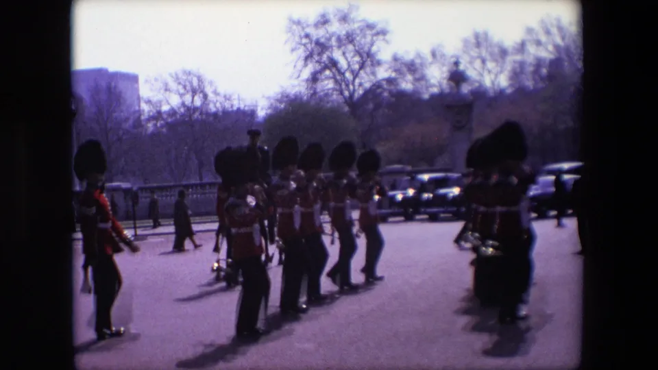 1967 London home-movie frame showing ceremonial Guards in bearskin hats moving in formation near pedestrians, parked or passing cars, trees, and stone roadside features.