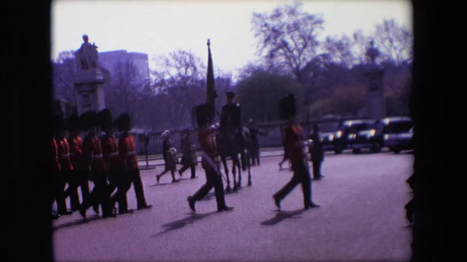 1967 London home-movie frame showing Guards in ceremonial formation with an upright flag or standard, a mounted officer, pedestrians, cars, trees, and a statue plinth nearby.