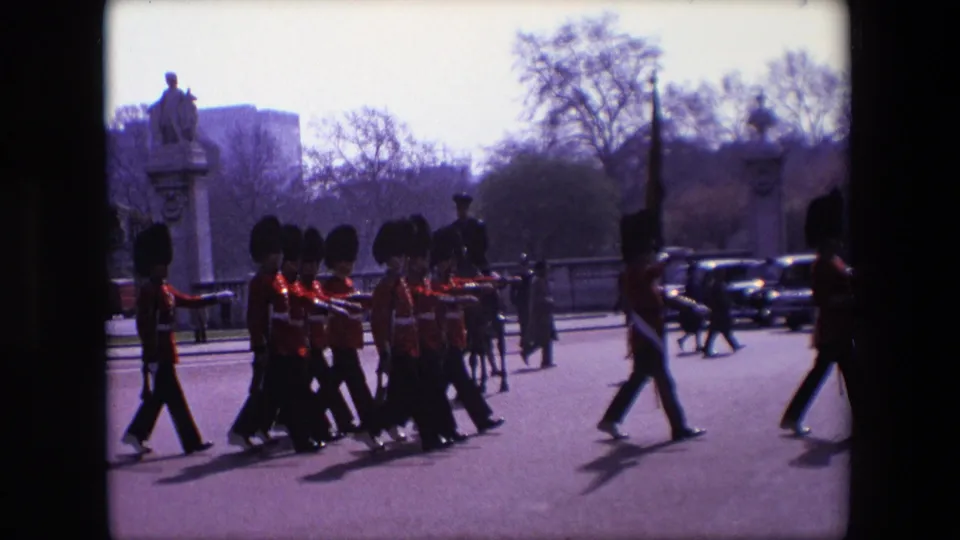 1967 London frame showing a compact group of ceremonial Guards in bearskin hats marching across the road, with pedestrians, dark cars, trees, and stonework behind them.