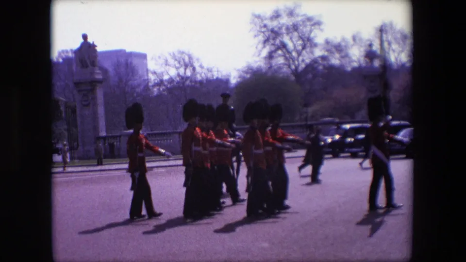 1967 London home-movie frame showing soldiers in red uniforms and tall dark headgear marching past spectators, classic cars, trees, and stone roadside features.