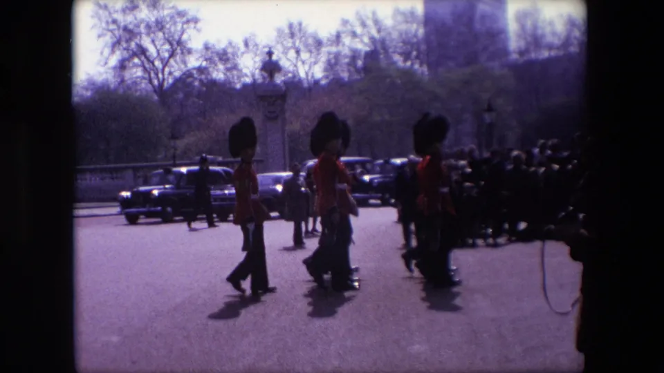 Home-movie frame from the supplied 1967 London clip showing guards marching in formation near a crowd of spectators, with classic cars and roadside trees visible.
