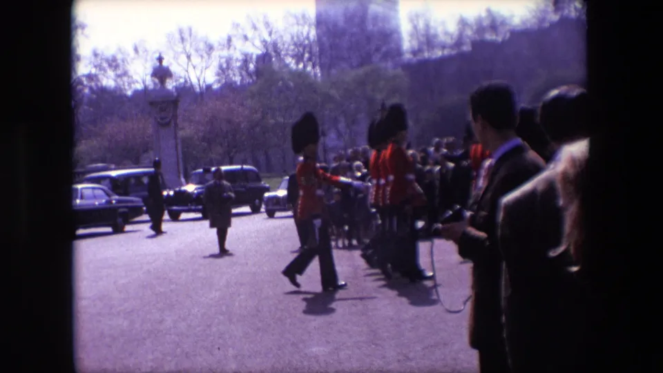 1967 London home-movie frame showing uniformed guards marching beside a line of spectators, with classic cars, stonework, trees, and a foreground bystander visible.