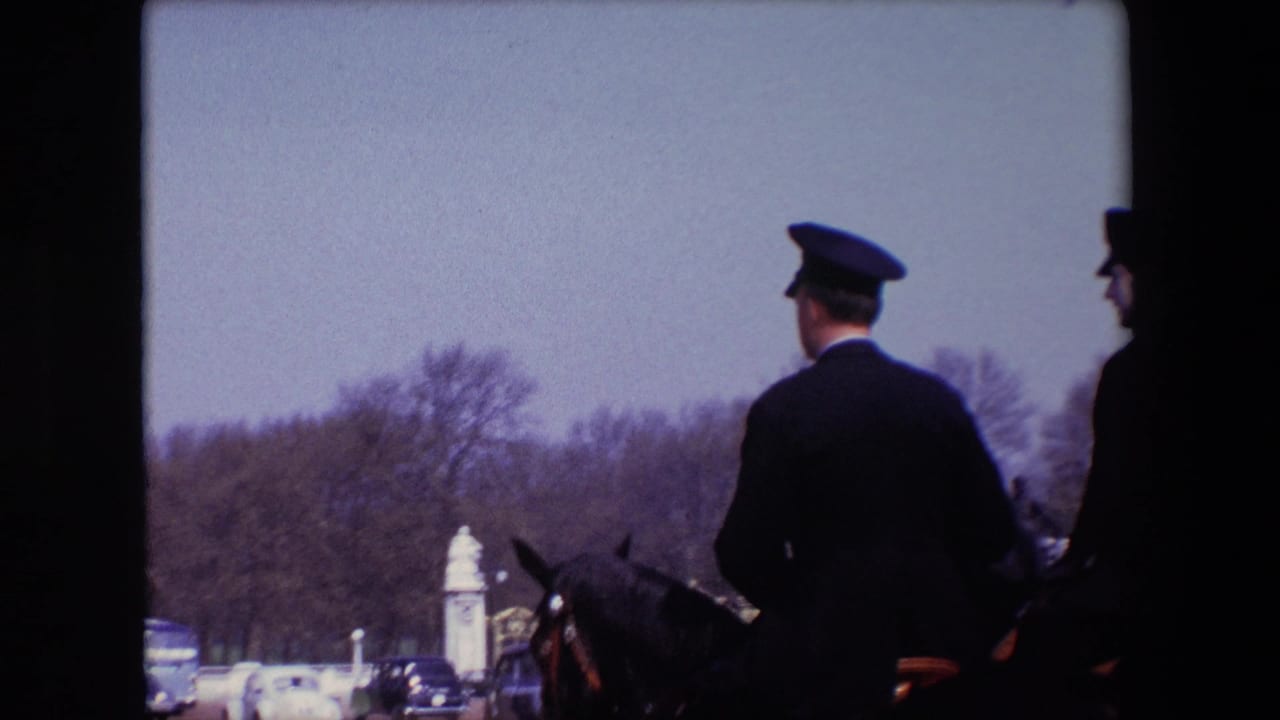 1967 London home-movie frame showing a mounted uniformed rider from behind, with cars, bare trees, and pale gate structures in the distance.