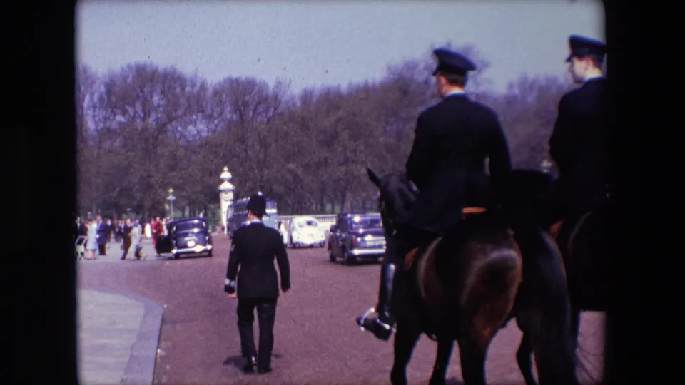 1967 London frame with mounted uniformed riders at the right edge, a uniformed figure walking ahead, pedestrians, cars, and tree-lined park space.