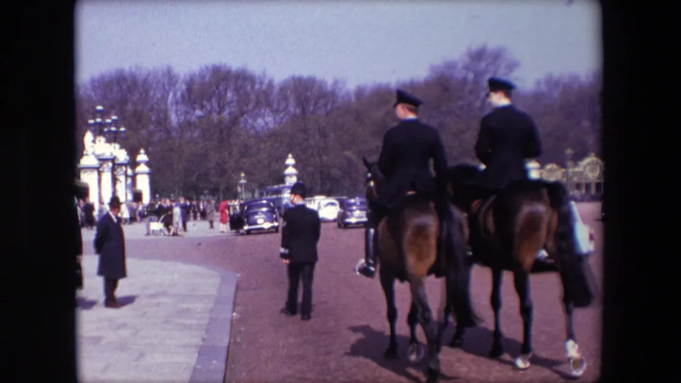 1967 London home-movie view of two mounted uniformed riders from behind, with pedestrians, period cars, sidewalk space, and pale gate structures along a tree-lined road.